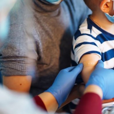 Gloved hands are smoothing a bandaid onto a child's upper arm. The child is on the lap of an adult carer.